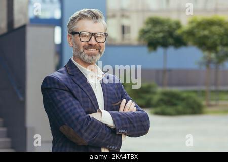 Ritratto dell'investitore grigio-haired maturo, uomo anziano negli occhiali che guardano la macchina fotografica e gesturing, uomo d'affari con le braccia incrociate fuori della costruzione dell'ufficio in vestito di affari, banchiere riuscito del boss. Foto Stock
