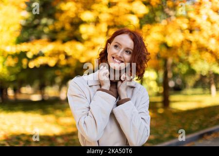 Felice giovane donna europea dai capelli rossi in impermeabile passeggiate nel parco cittadino, guardare la copia spazio in autunno, primo piano Foto Stock