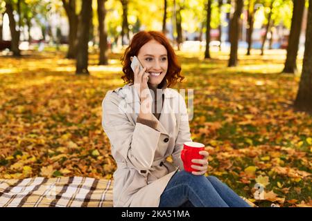 Allegra giovane donna europea dai capelli rossi in impermeabile gode di una tazza di bevanda, chiamate per telefono nel parco cittadino Foto Stock