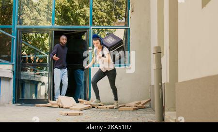 Persona che consegna il fastfood che cade le scatole della pizza impilata alla porta anteriore del cliente, inciampando e correndo. Cattivo servizio di consegna, pizzeria takeaway dipendente cercando di catturare i pacchi in caduta. Scatto con cavalletto. Foto Stock