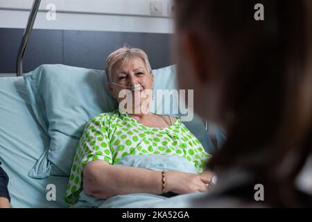 Donna anziana che parla con una ragazza irriconoscibile. Primo piano della felice nonna visitata dalla nipote nella stanza dell'ospedale geriatrico. Donna sorridente che parla con una parente femminile nel letto del sanatorio. Foto Stock