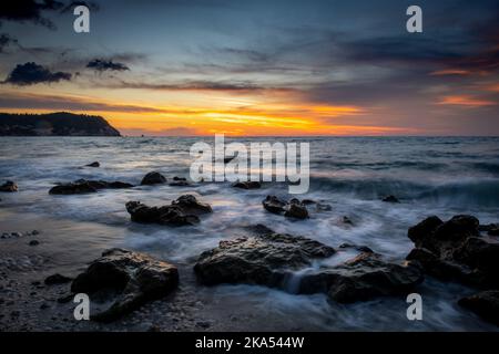 Una lunga esposizione di mare di onde che si lavano sopra le rocce sulla spiaggia al tramonto. Foto Stock
