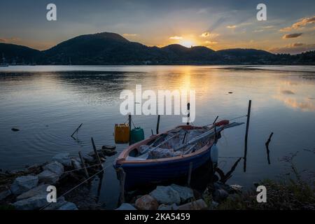 Una vecchia barca di legno su una baia di mare con acque calme con il sole che sorge sulla cima della collina. Foto Stock