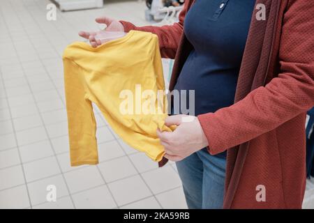 Donna incinta sceglie un vestito giallo - corpetto per un bambino in un negozio per acquistare Foto Stock