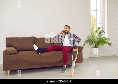Uomo felice con gamba rotta che parla con qualcuno al telefono mentre si siede sul divano a casa Foto Stock