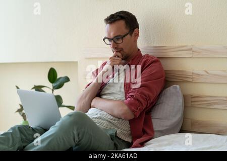 Portato via l'investitore europeo di uomo lavori si siede in camera da letto sul letto tiene traccia delle quotazioni azionarie tramite laptop Foto Stock