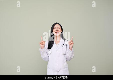 Stupito e allegro giovane medico asiatico femminile in uniforme e stetoscopio guardando verso l'alto, puntando le dita verso l'alto, in piedi isolato sfondo grigio. Promozione speciale Foto Stock