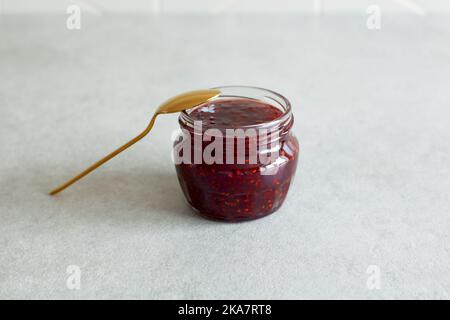 Pronto a mangiare confettura di lamponi in vaso di vetro aperto con cucchiaio d'oro isolato sul tavolo da cucina in pietra grigia. Ripresa ad angolo, messa a fuoco selettiva. Foto Stock