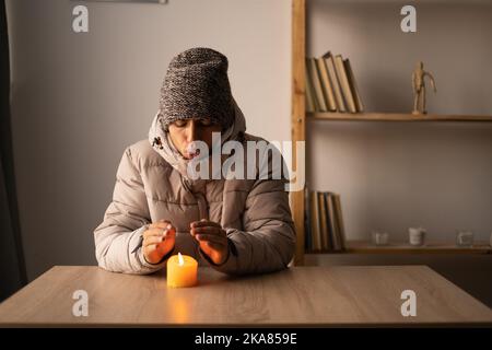 Il congelamento del giovane uomo nei vestiti di inverno riscalda le mani sulla candela che brucia. Arresto del riscaldamento e dell'elettricità, interruzione dell'alimentazione, blackout, scarico del carico o. Foto Stock