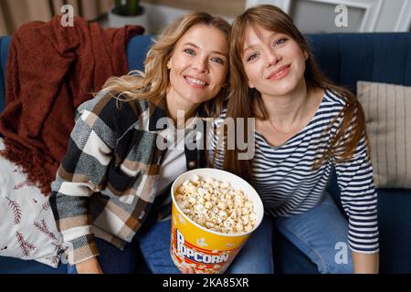 Ritratto di madre matura e figlia adulta seduta sul divano con secchio di popcorn mentre guarda il film Foto Stock