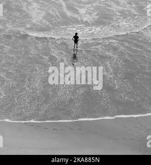 Una scala di grigi verticale di una persona in piedi sulla sabbia della spiaggia contro la superficie dell'acqua. Foto Stock