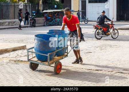 Un cubano afro-caraibico spinge un carrello con due serbatoi di plastica. È visto su una strada di ciottoli della città. Foto Stock
