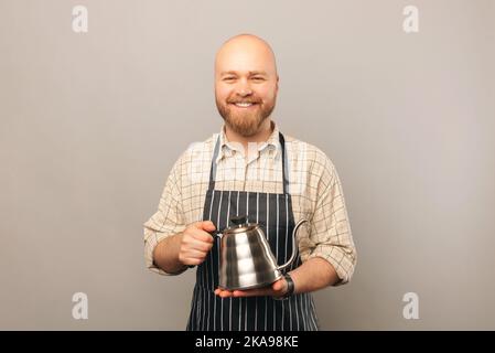 Il barista sorridente è in possesso di un bollitore per caffè all'americana in metallo per preparare il caffè. Studio girato su sfondo grigio. Foto Stock