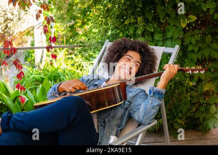 Bel giovane afro-italiano uomo di corsa mista in abbigliamento casual suonare la chitarra in giardino. Un ragazzo multirazziale suona uno strumento musicale riposa e si rilassa Foto Stock