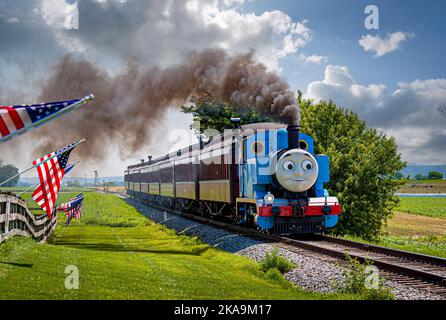 Strasburg, Pennsylvania. 48 2021 Giugno - Vista di Thomas il treno che tira le vetture del passeggero mentre passa le bandiere americane su un recinto in un giorno di sole Foto Stock