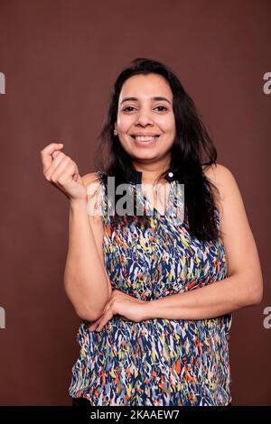 Sorridente donna indiana allegra ritratto ridente, donna felice che tiene la mano piegata, schiaffo dita. Persona spensierata che posa la vista anteriore studio medio girato su sfondo marrone Foto Stock