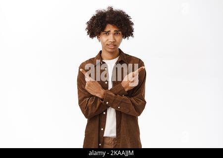 Ritratto del ragazzo di gen z sorridente con capelli afro, puntando le dita lateralmente, mostrando il lato sinistro e destro, logo o banner di prodotto, dare la scelta, in piedi Foto Stock