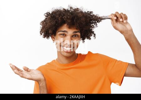 Primo piano ritratto di giovane uomo che tocca i ricci, ragazzo con capelli ricci sorridente e ridente, in piedi su sfondo bianco in t-shirt arancione Foto Stock