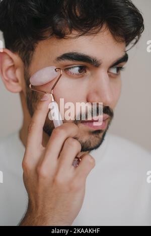 Primo piano ritratto di un uomo che guarda lontano e che fa massaggio linfatico del viso con rullo di giada per sollevare la pelle isolata su sfondo bianco. Routine di bellezza Foto Stock