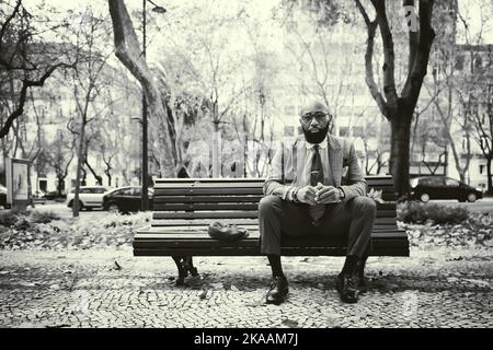 Foto in bianco e nero con un elegante imprenditore africano balduto in un abito formale sartoriale alla moda con cravatta e occhiali seduti Foto Stock