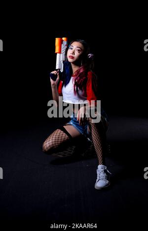 Giovane donna che tiene la pistola d'acqua vestita in costume di Harlequin su una strada di Asphalt Foto Stock