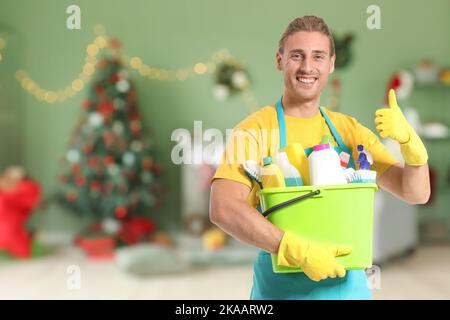 Janitor maschio con i rifornimenti di pulizia che mostrano il gesto pollice-in su nella stanza alla vigilia di Natale Foto Stock