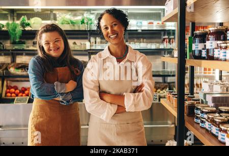 I lavoratori di successo del negozio di alimentari sorridono alla macchina fotografica mentre si trovano in piedi insieme nel loro negozio. Donna felice con sindrome di Down che lavora in un locale superm Foto Stock