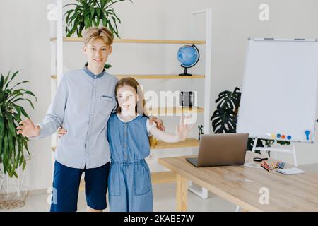 Ragazza e ragazzo adolescenti in piedi accanto al tavolo con un computer portatile in classe che guarda alla fotocamera. Foto Stock