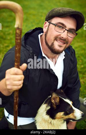 Solo un altro giorno in fattoria. Ritratto di un giovane contadino allegro e il suo cane che si rilassa su un campo verde in una fattoria durante il giorno. Foto Stock