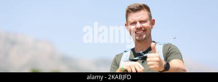 Un uomo sorridente che tiene i pollici in su tiene i bastoni scandinavi e si leva in piedi sulla montagna Foto Stock