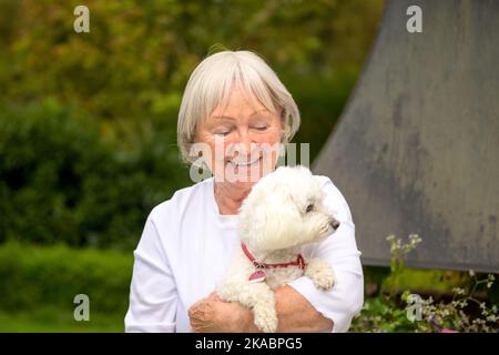 Anziana signora dai capelli grigi tiene amorevolmente il suo carino cane bianco tra le braccia Foto Stock