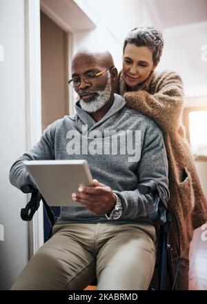 Guarda alcuni dei nostri video preferiti. Una coppia affettuosa con un tablet a casa con l'uomo seduto su una sedia a rotelle. Foto Stock