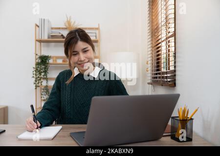 studentessa asiatica che studia a distanza da casa, utilizzando un computer portatile, prendendo appunti su blocco note durante la lezione online, il concetto di e-learning, sorridendo Foto Stock