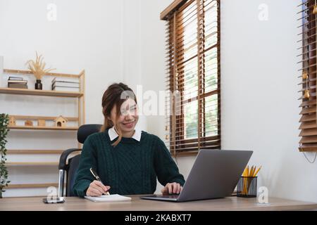 studentessa asiatica che studia a distanza da casa, utilizzando un computer portatile, prendendo appunti su blocco note durante la lezione online, il concetto di e-learning, sorridendo Foto Stock