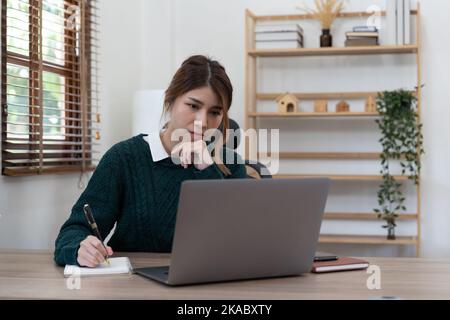 Concentrate le studentesse asiatiche studiando a distanza da casa, utilizzando un computer portatile, prendendo appunti sul blocco note durante la lezione online, il concetto di e-learning Foto Stock