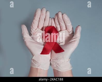 Arco a nastro rosso per la consapevolezza dell'AIDS e dell'HIV, in primo piano con la mano del medico. Foto Stock