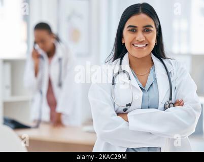 Donna indiana, dottore e braccia attraversate in ospedale con fiducia, obiettivi di benessere o motivazione sanitaria. Ritratto, sorriso o felice operatore medico, vita Foto Stock