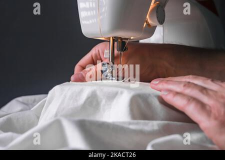 Mano maschile alla macchina da cucire durante il processo di lavorazione con primo piano in tessuto di cotone naturale. Foto Stock