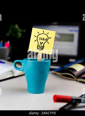 Un leggero scarabocchio di lampadina disegnato su un foglietto appiccicoso all'interno di una tazza di caffè alla scrivania dell'ufficio. Idee, ispirazione e pensiero creativo vengono con una tazza di mattina di Foto Stock