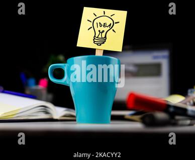Un leggero scarabocchio di lampadina disegnato su un foglietto appiccicoso all'interno di una tazza di caffè alla scrivania dell'ufficio. Idee, ispirazione e pensiero creativo vengono con una tazza di mattina di Foto Stock