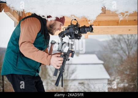 Uomo felice del videografo che scatta le riprese, usando la macchina fotografica montata sull'apparecchiatura di stabilizzatore di gimbal in inverno. Foto Stock