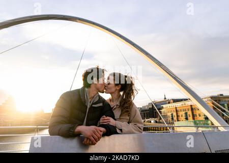 Un'ampia vista di una giovane coppia che si trova fianco a fianco condividendo un bacio insieme su un ponte della città, sono innamorati. Foto Stock