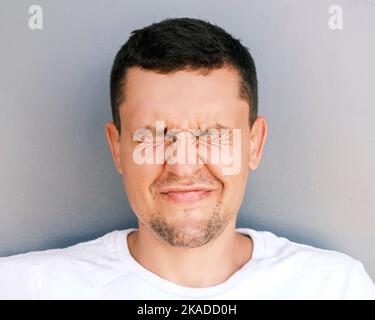 Ritratto di un uomo divertente con stoppia chiudere gli occhi e sorridere su uno sfondo chiaro. Un ragazzo emozionale in una T-shirt bianca si scinde Foto Stock