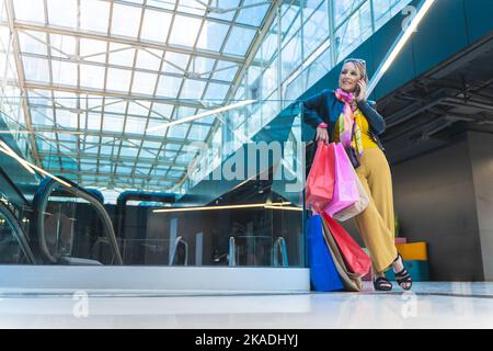 Donna matura che parla smartphone con borse shopping nel centro commerciale. Foto Stock