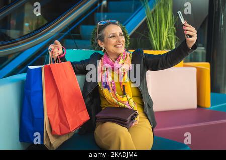 Signora con le borse della spesa seduti e prendere un selfie in un centro commerciale. Felice donna anziana sorridente che riposa dopo il giorno del negozio. Foto Stock