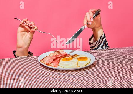 Le mani femminili con forchetta e coltello mangiano la colazione inglese con uova fritte e pancetta Foto Stock