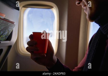 Sorridente passeggero della compagnia aerea con una tazza di caffè di carta guardando avanti nella finestra dell'aereo. L'uomo beve il tè a bordo. Il maschio trattiene bevande o acqua durante Foto Stock