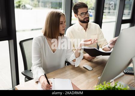 Uomo d'affari che tiene la cartella di carta vicino al nuovo lavoratore che scrive sul notebook e sul computer in ufficio, immagine di scorta Foto Stock