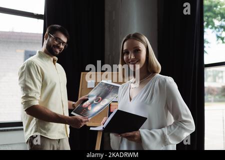 Sorridente intern tenendo il notebook e guardando la fotocamera vicino a un uomo d'affari con grafici in ufficio, immagine stock Foto Stock