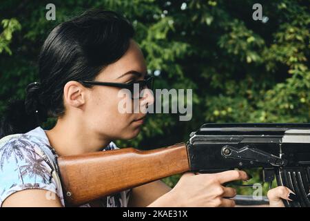 Una giovane ragazza in occhiali da sole scuri spara da una mitragliatrice e alberi verdi Foto Stock